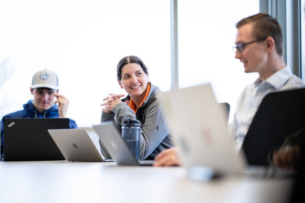 Three people sit chatting to each other. Three laptops are visible and one is a blue hoodie and a  cap branded GV is looking at his laptop.